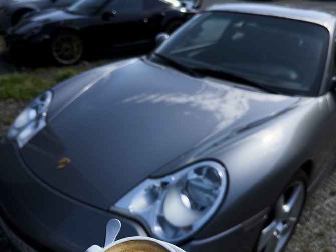 Eine Tasse Cappuccino vor einem grauen Sportwagen auf einem Parkplatz in den Bergen