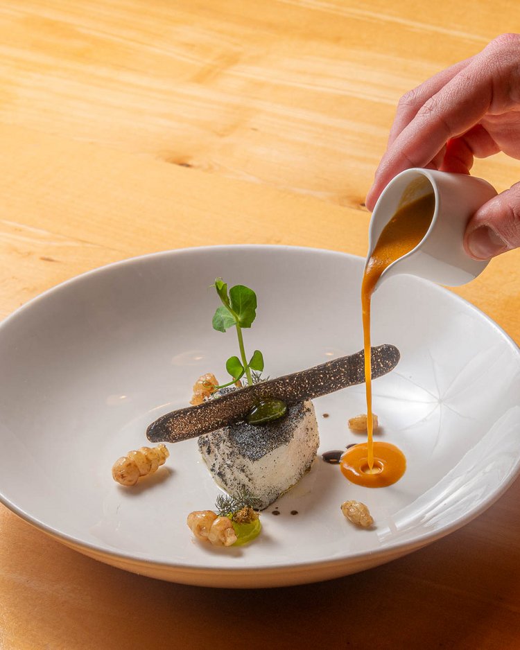 Hand gießt Sauce auf ein kunstvoll angerichtetes Gericht im weißen Teller