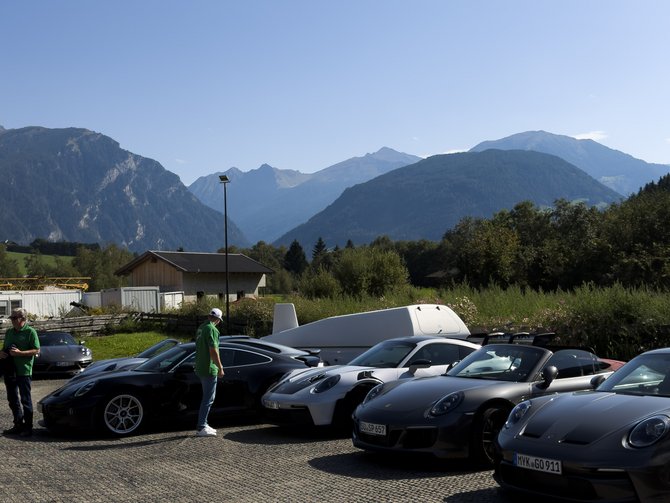 Mehrere Sportwagen auf einem Parkplatz vor einer Bergkulisse, zwei Personen stehen neben den Autos
