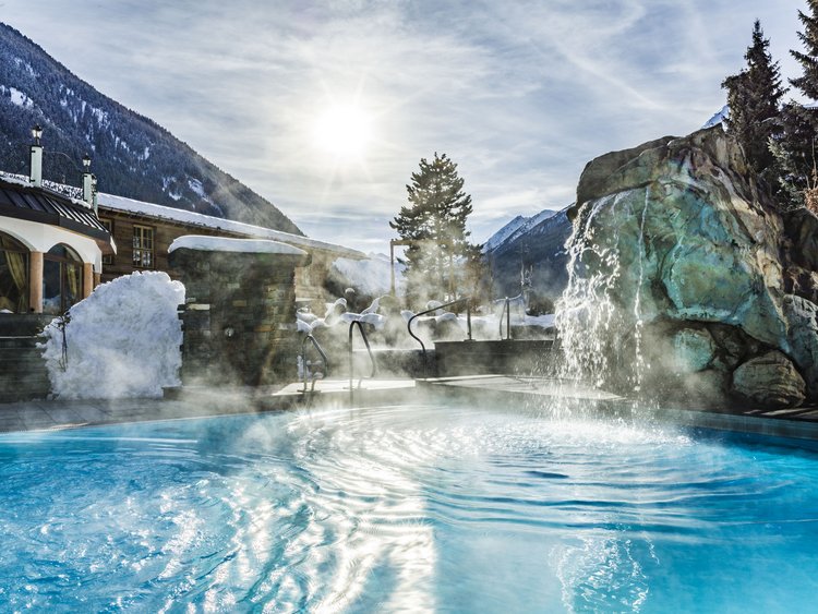 Outdoor hot tub with waterfall in snowy mountain landscape under bright sun