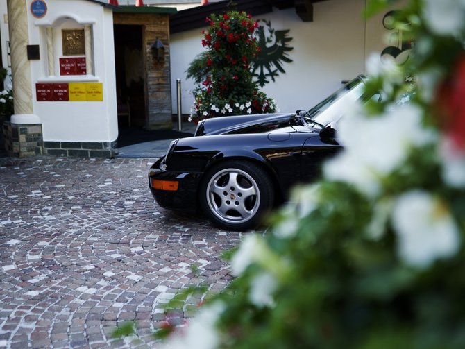 Schwarzer Sportwagen vor einem Gebäude auf einem gepflasterten Hof, dahinter bewaldete Berge