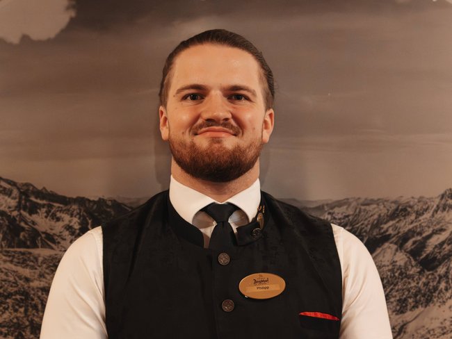 Discover the Jagdhof, Stubaital, and Tyrol Man in traditional vest in front of wooden frame with Jagdhof logo and mountain landscape
