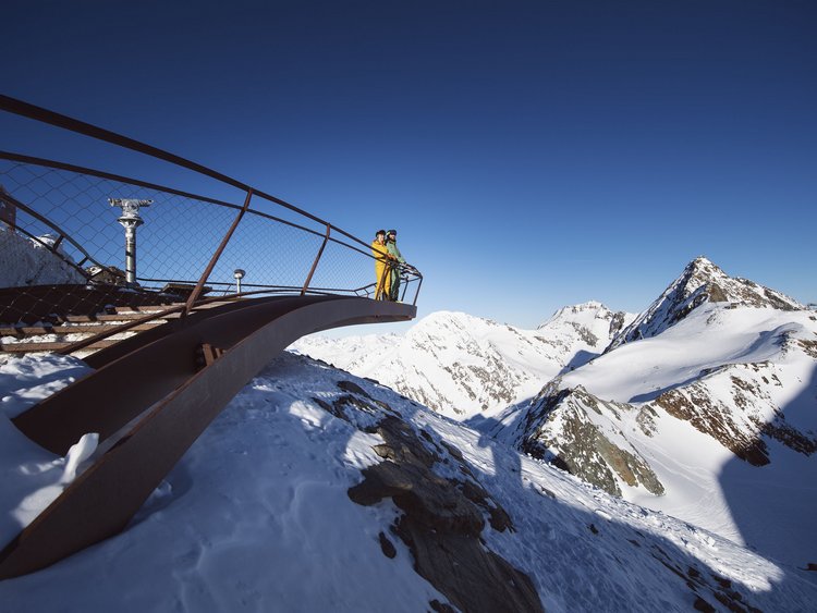 Vacanze attive in hotel: l’Austria in montagna Due persone su una piattaforma panoramica sopra le montagne innevate sotto un cielo limpido