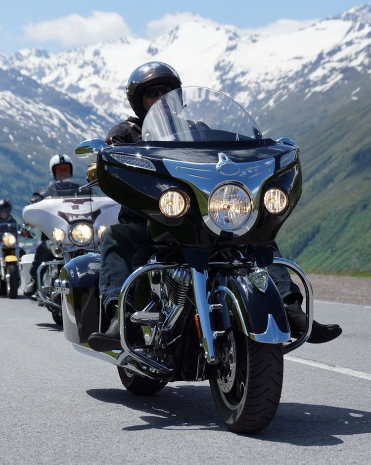 Gruppe von Motorradfahrern fährt auf Bergstraße mit schneebedeckten Gipfeln im Hintergrund