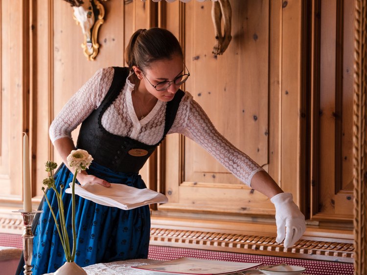 Discover the Jagdhof, Stubaital, and Tyrol Waitress in traditional dress setting table in wood-paneled restaurant with hunting trophies