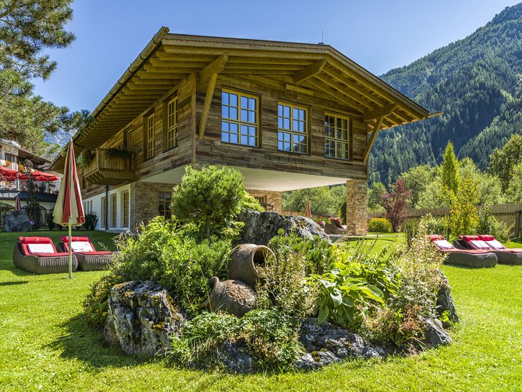 Holzhaus auf Stelzen mit Garten und Liegestühlen vor Bergkulisse